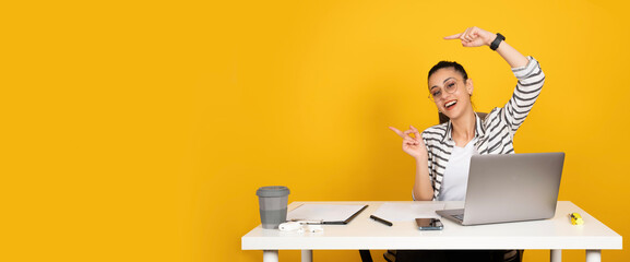 Woman pointing copy space, young happy cheerful girl wear casual shirt sit at office desk woman pointing copy space. Using laptop fingers showing aside mock up area. Isolated yellow background. 
