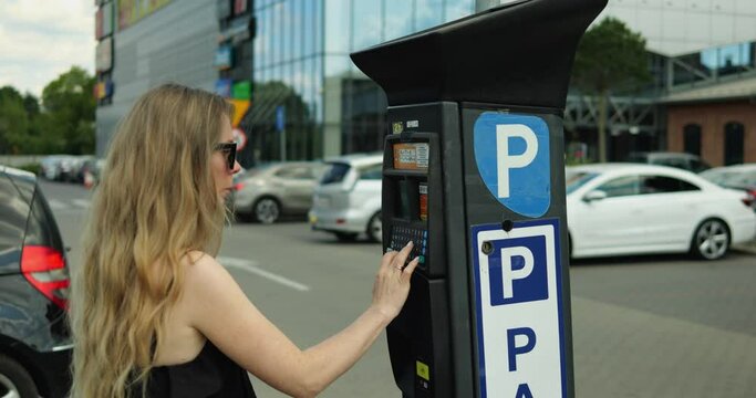 Woman in sunglasses and dress type registration plate number on parking meter. Elegant generation z female driver press keypad buttons on parking kiosk to buy parking ticket.