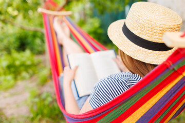 Young woman reading book while lying in comfortable hammock at green garden