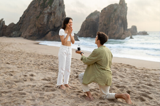 Full length shot of loving man with engagement ring proposing to surprised lady, guy standing on knee on ocean beach