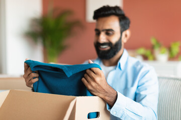 Glad contented Indian buyer man unpacking box with clothes indoor