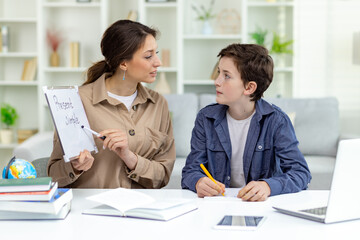 Home education, woman teaches boy at home sitting at table in living room, mother explains...
