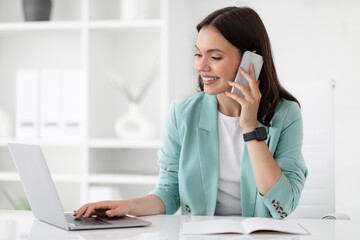 Cheerful millennial caucasian lady in suit typing on computer, calls by smartphone at workplace
