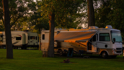 Rv and trailers parked at campsites amongst tall trees in early morning with grass and sunrise...