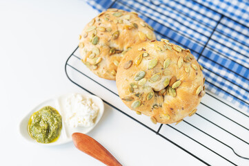 A freshly baked bagel with sunflower seeds lies on a metal stand along with a plate of cheese and pesto sauce and a wooden knife.