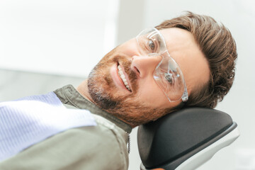 Handsome smiling bearded man in protective glasses sitting in dental chair, looking at camera
