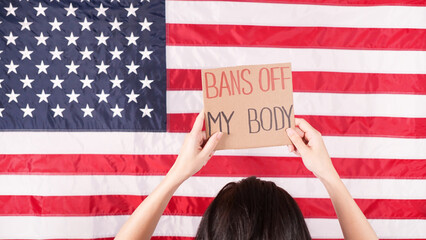 Young woman protester holds cardboard with Bans Of My Body signs against USA flag on background....