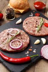 Raw beef hamburger patties on stone plate, vegetables and spices, wooden background.