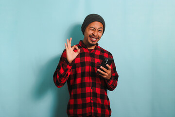 Satisfied Young Asian man with a beanie hat and a red plaid flannel shirt is winking his eyes while holding a mobile phone and showing an OK gesture, isolated on a blue background - obrazy, fototapety, plakaty