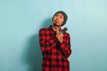 Pensive young Asian man with a beanie hat and red plaid flannel shirt thinking about what movie to watch while holding a TV remote, isolated on a blue background