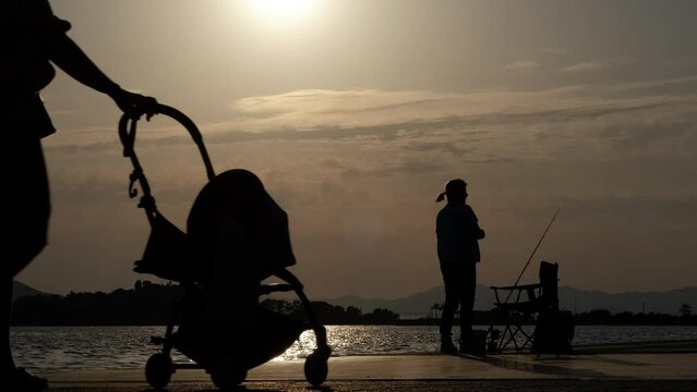 City beach silhouette with fishingman hobby. A silhouette of female with fishing rob and people relaxing on the sunset beach during summer holidays.