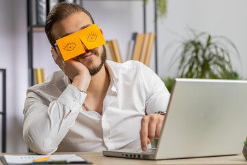 Inefficient tired young businessman working sleeping on laptop computer with eyes stickers on face...