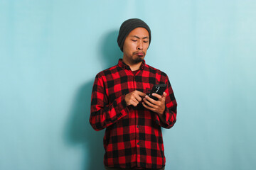 An upset Young Asian man with a beanie hat and a red plaid flannel shirt is holding a mobile phone, experiencing problems, and feeling angry and annoyed, isolated on a blue background
