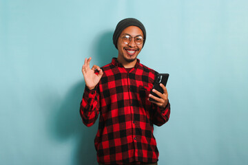 Satisfied young Asian man wearing eyeglasses, a beanie hat, and a red plaid flannel shirt is using a smartphone and showing an OK gesture, isolated on a blue background