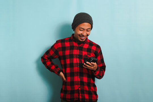 Happy young Asian man with a beanie hat and a red plaid flannel shirt is using a smartphone to check social media and shop online, isolated on a blue background