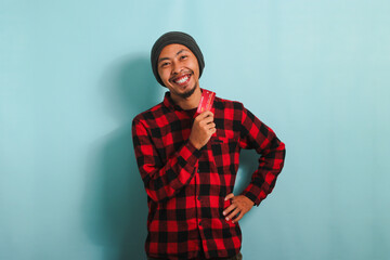 Fototapeta premium Excited young Asian man with a beanie hat and red plaid flannel shirt is holding a bank credit card in his hands, ready to make a purchase in an online store while standing against a blue background
