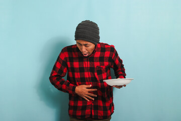 Young Asian man with a beanie hat and a red plaid flannel shirt is feeling nauseous while holding...