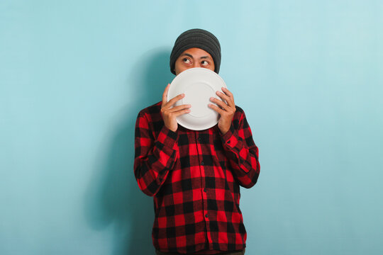 Hungry Young Asian man with a beanie hat and a red plaid flannel shirt is looking aside, thinking about what to eat, while holding an empty white plate in his hand, isolated on a blue background