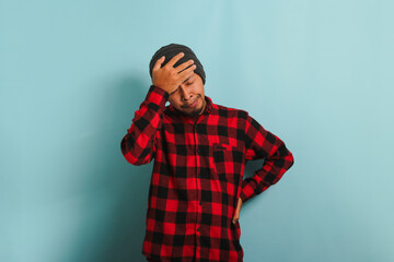 A young Asian man with a beanie hat and a red plaid flannel shirt is suffering from a headache, feeling desperate and stressed due to the pain and migraine, while standing against a blue background