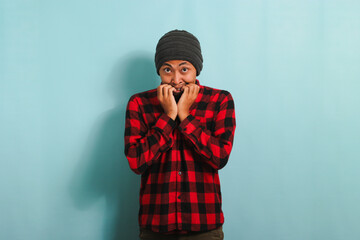 Embarrassed young Asian man with beanie hat and red plaid flannel shirt is shocked, covering his mouth in response to a mistake and feeling bad for saying something wrong, isolated on blue background