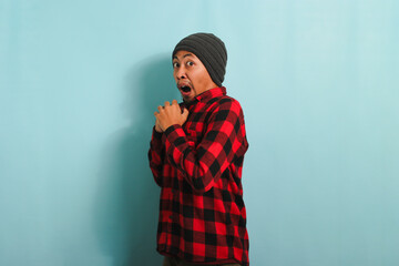 Scared Young Asian man with a beanie hat and red plaid flannel shirt has an open mouth, looking surprised and shocked, reacting to something horrible, isolated on a blue background