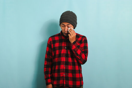 A crying young Asian man with a beanie hat and a red plaid flannel shirt wipes tears with a tissue while standing against a blue background
