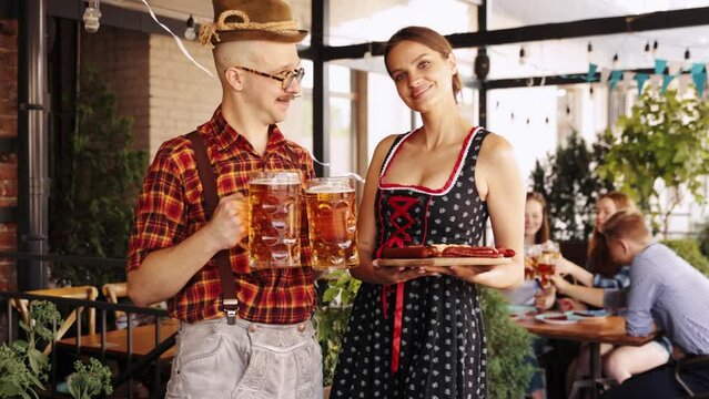Man and woman in traditional clothes standing with lager beer and appetizers at pub. Serving beer to group of friends. Concept of oktoberfest, traditional taste, friendship, leisure time, enjoyment