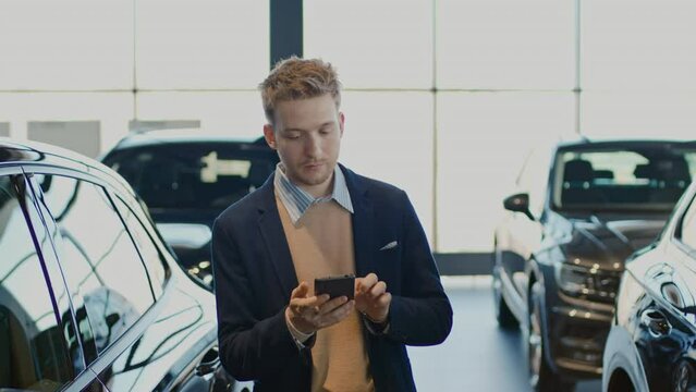 Medium shot of young Caucasian male customer in smart casual jacket and cashmere sweater walking around car dealership, looking at smartphone and checking out new models on display