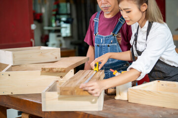 Senior male carpenter teaching young woman to use equipment standing with bearded craftsman improving DIY furniture. Diverse ethnic family in carpentry while working at carpentry workshop.