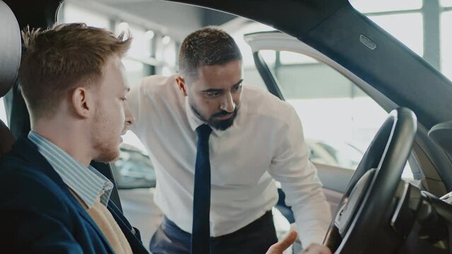 Medium close-up shot of Caucasian male customer sitting in driver seat of new car in dealership, holding steering wheel, biracial sales manager in white shirt and tie demonstrating vehicle features