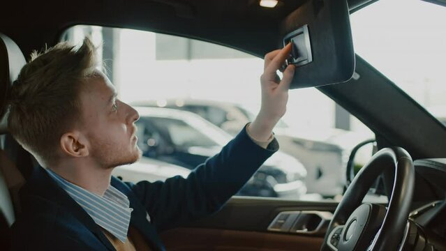 Medium shot of young Caucasian male buyer sitting in driver seat of new car in dealership, touching steering wheel, adjusting his red hair in mirror on windshield visor, opening storage compartment