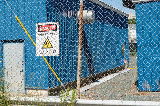 A danger sign, high voltage, with keep out electricity symbol hanging on a mesh wire fence with two blue buildings in an enclosed yard. The electrical station storage building has a single grey door. 