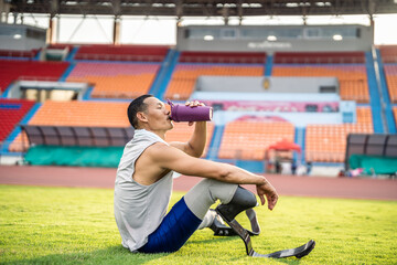 Asian para-athlete with prosthetic blades drinking water in stadium.