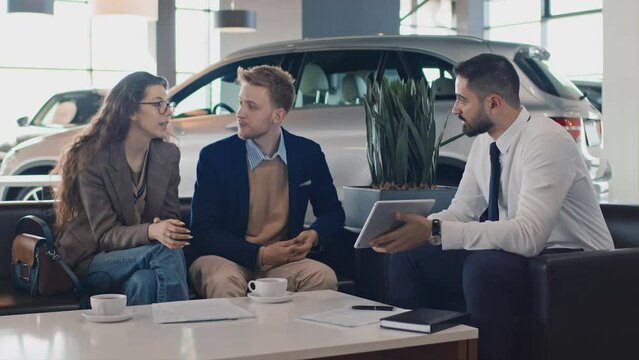 Medium shot of young Caucasian husband and wife sitting in car showroom, Middle Eastern salesman calculating loan conditions and monthly payments on tablet computer, and negotiating purchase terms