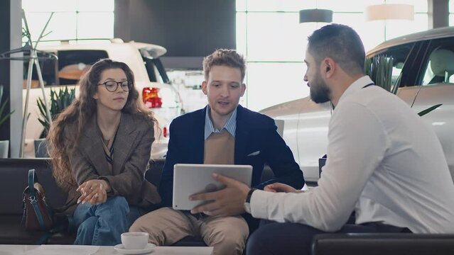 Medium shot of young Caucasian couple sitting on couch in car dealership, Arab male sales manager showing available car colours on tablet computer, buyers discussing, smiling and nodding
