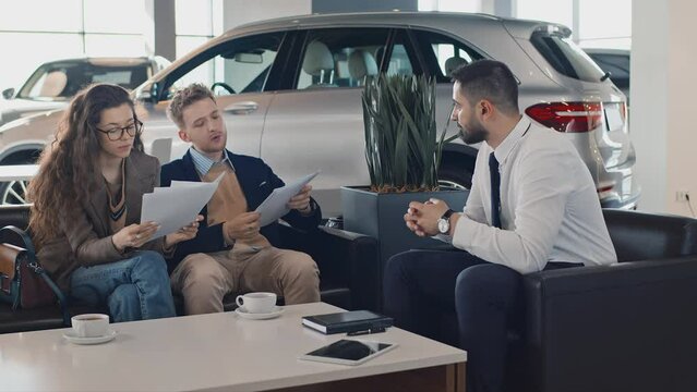 Full shot of young Caucasian husband and wife sitting on couch in dealership, reading car loan contract, man asking sales manager for clarification of financial termsFull shot of young Caucasian husba