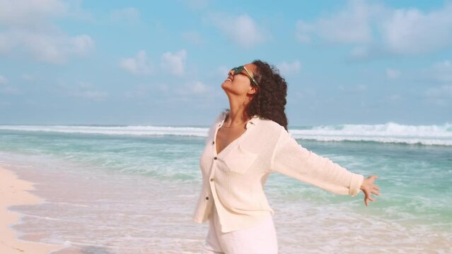 Young cheerful Indian woman in sunglasses and light clothing is twirling stands on beach rejoicing at good weather and opportunity to visit seaside resorts on summer vacation located on ocean