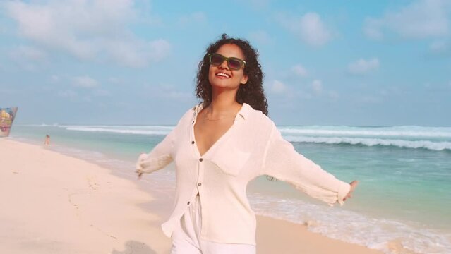 Young cheerful sympathetic Indian woman brunette experiences positive emotions and spins stands on sandy sea coast with picturesque view of ocean located on beach near sea. Weekend, holidays, vacation