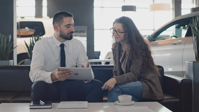 Medium shot of young Caucasian female customer sitting in auto dealership, listening to salesman with tablet demonstrating available car models, discussing features and options, pointing to display