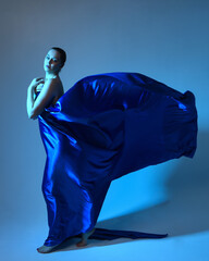 Full length portrait of beautiful female model wearing elegant flowing toga gown of blue silk drapery, isolated on studio background with cinematic colourful lighting.