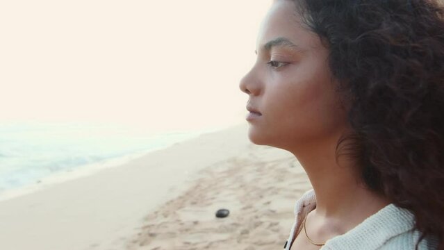 Young thoughtful Indian woman looks into distance and thinks about plans for future and smiles slightly imagining another trip or successful career stands on tropical beach near sea.