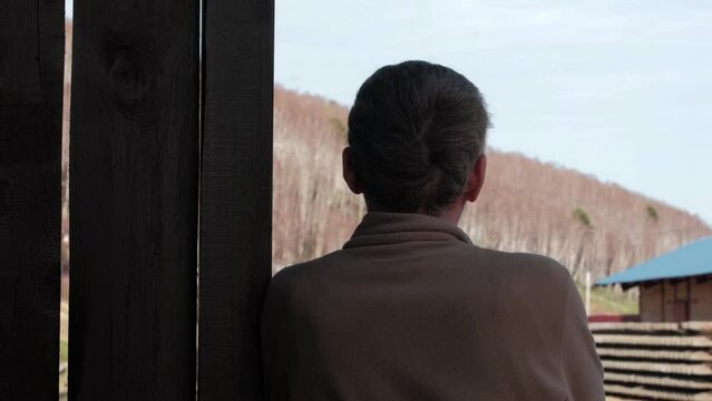 middle aged man with glasses approaches and leans against a wooden wall at an old railway station. he looks around and admires the landscape. rear view from the back.