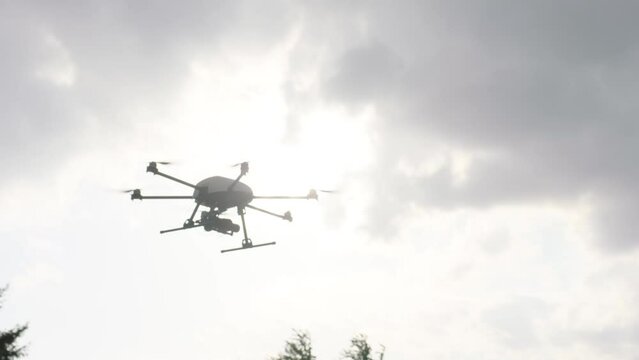 Handheld camera tracks a white drone with six propellers, flying towards the right in a cloudy sky with the sun peeping through