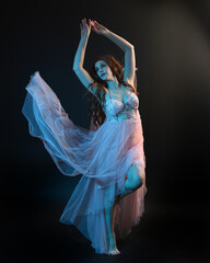 Fototapeta premium Full length portrait of beautiful brunette woman dancer, wearing ethereal gown, dancing with flowing fabric. isolated on dark studio background with cinematic colourful lighting.