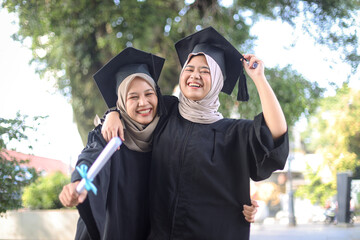 Two young muslim ladies in graduation costumes posing at camera at university campus, holding...