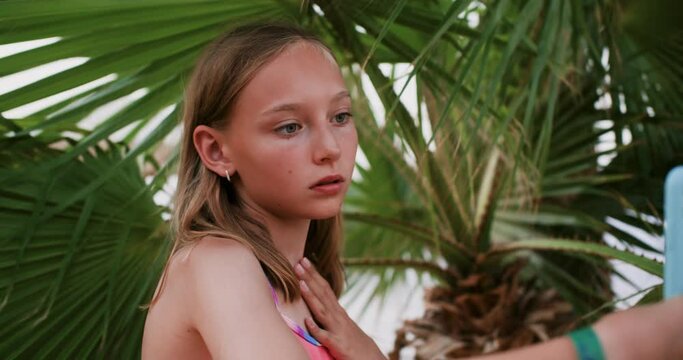 Model blogger blonde teen girl makes selfie on smartphone under palm tree on beach resting in tropics on vacations. Teenager photographing herself for social media. Travel, tourism, creating content.