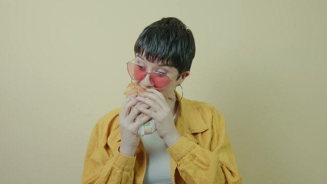 Modern short-haired woman voraciously chomping a burger in a studio background. Slow motion