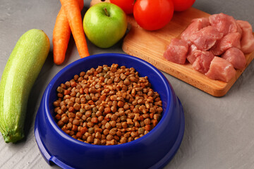 Bowl with dry pet food, raw meat and natural products on color wooden background, closeup