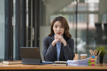 An attractive beautiful millennial Asian businesswoman or female marketing manager sits at her desk with hands on her chin, smiling and looking at the camera.
