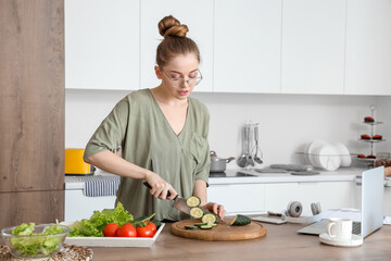 Beautiful young woman cutting cucumber at table with modern laptop in light kitchen